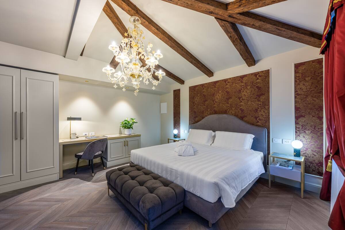 Une chambre à coucher avec un grand lit, un bureau et une chaise. Un lustre est suspendu au-dessus du lit, et il y a une table de chevet avec une lampe.