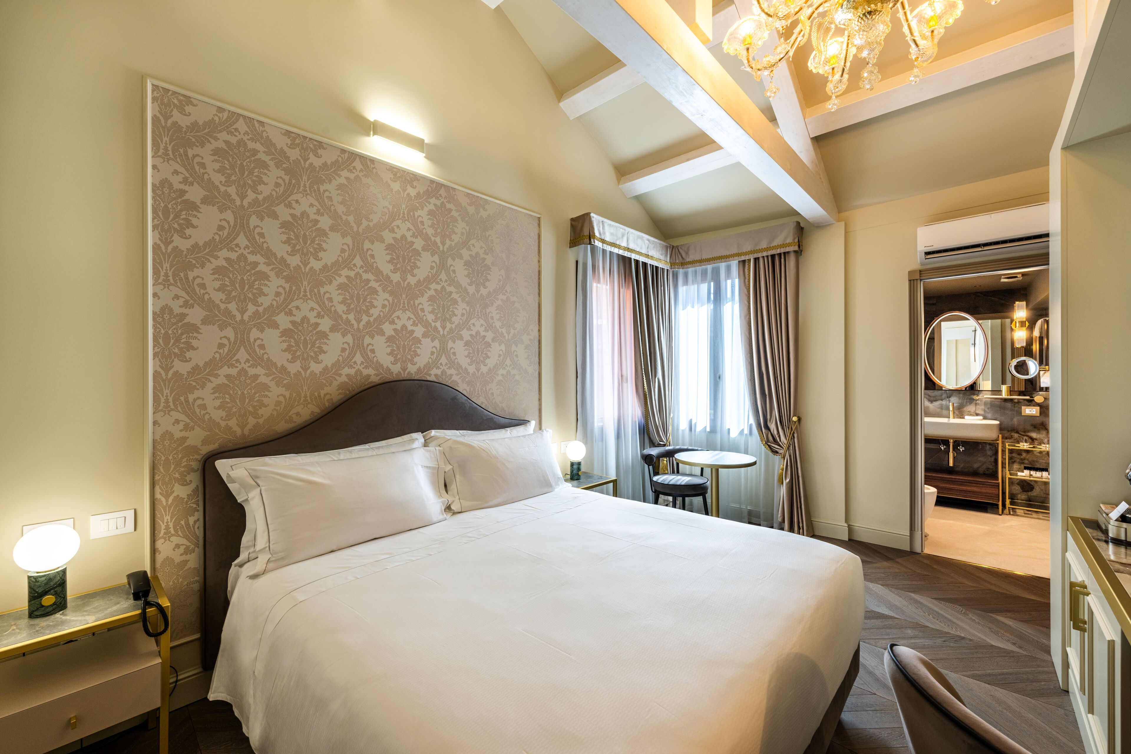 Quarto de hotel com uma cama grande, mesa de cabeceira com abajur, uma cadeira, mesa redonda pequena e vista para o banheiro.