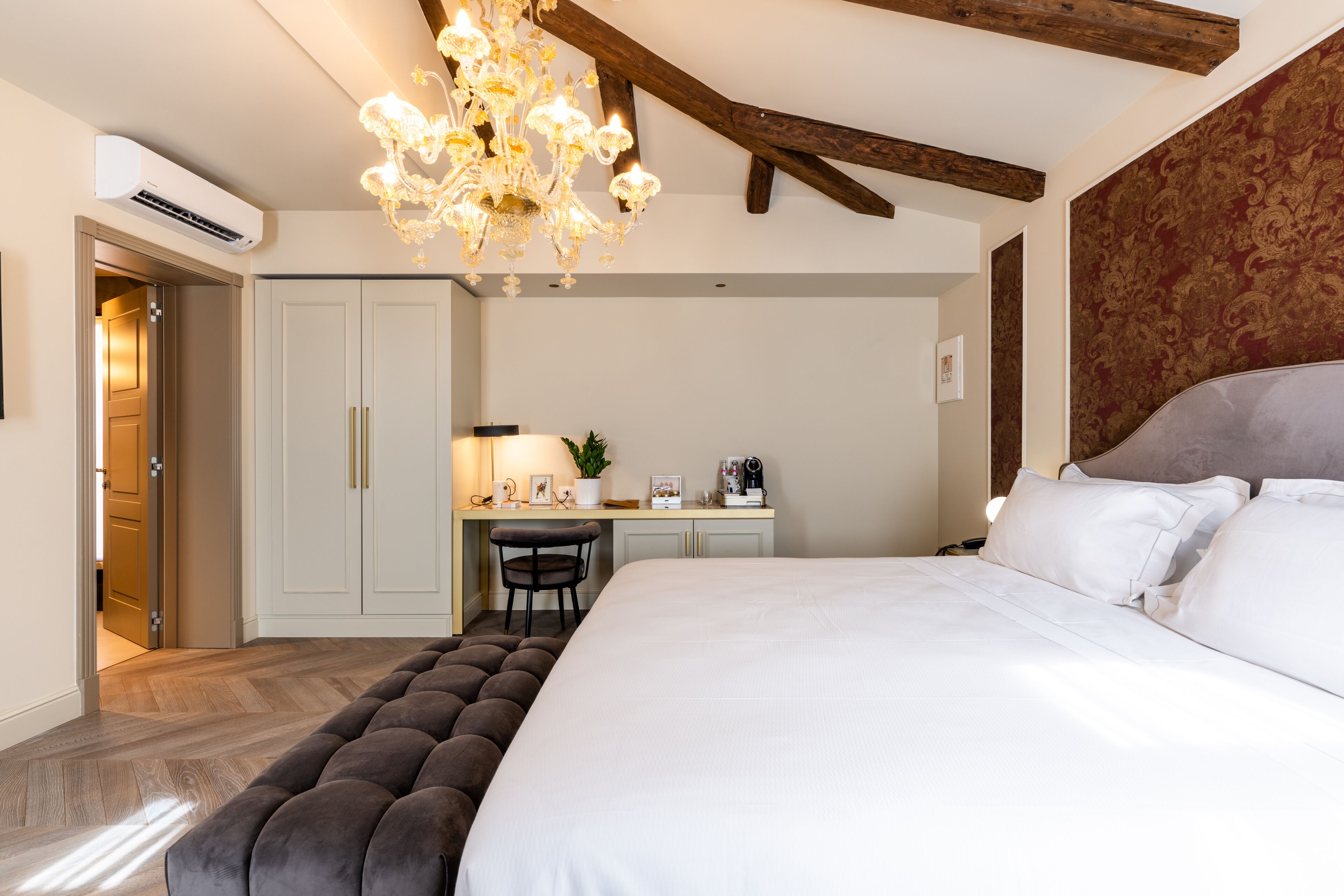 A bedroom with a large bed, a desk, and a chair. There is a chandelier above the bed and an air conditioning unit on the wall.