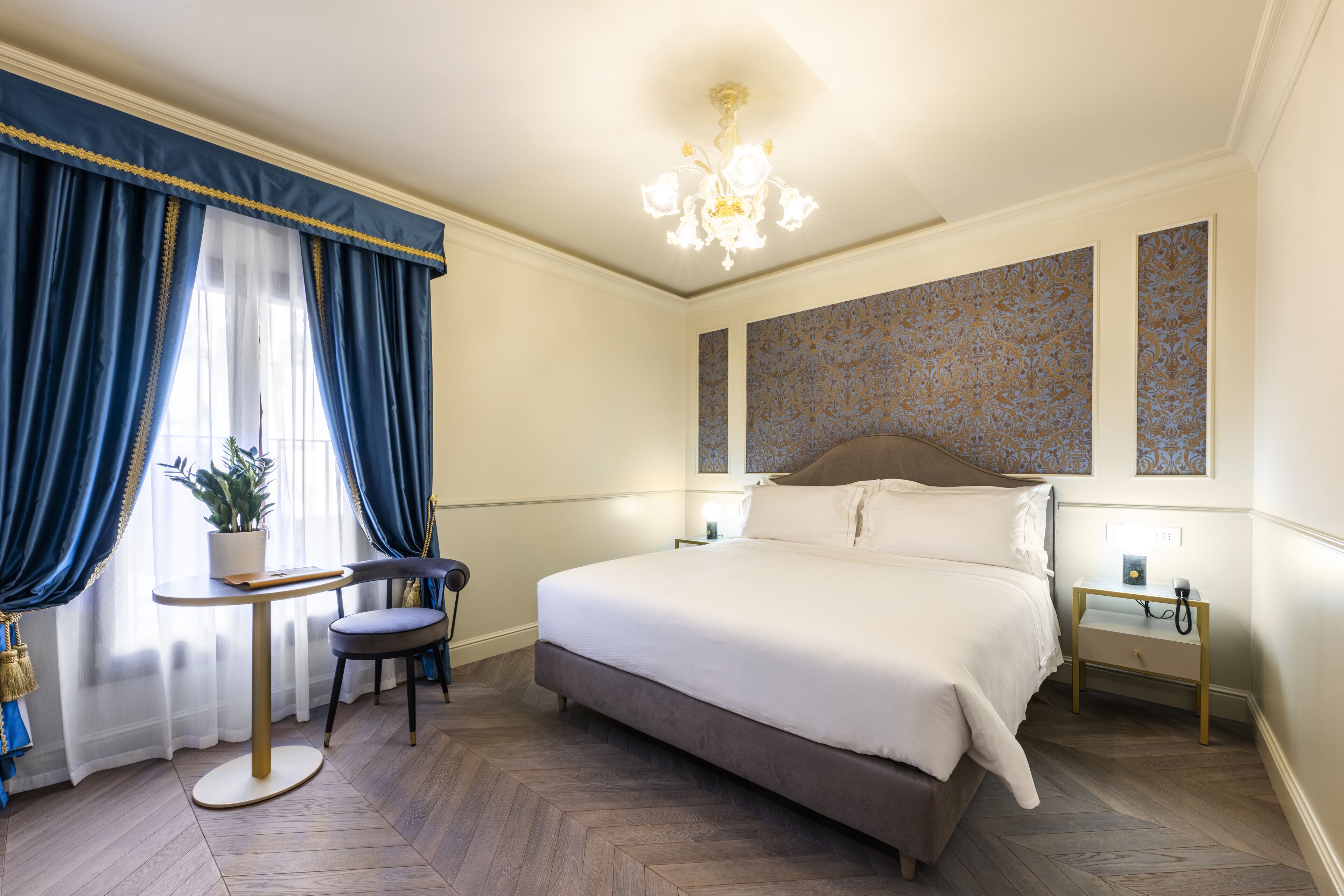 A hotel room with a large bed, a small table with a chair, a potted plant, and a nightstand with a lamp.
