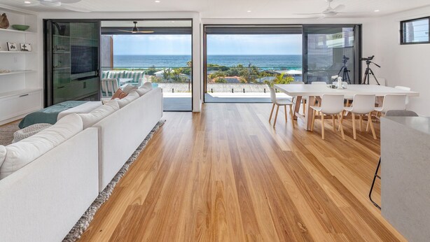 Ein modernes Wohnzimmer mit Holzboden, weißen Möbeln und einer großen Schiebeglastür, die auf einen Balkon mit Meerblick führt.