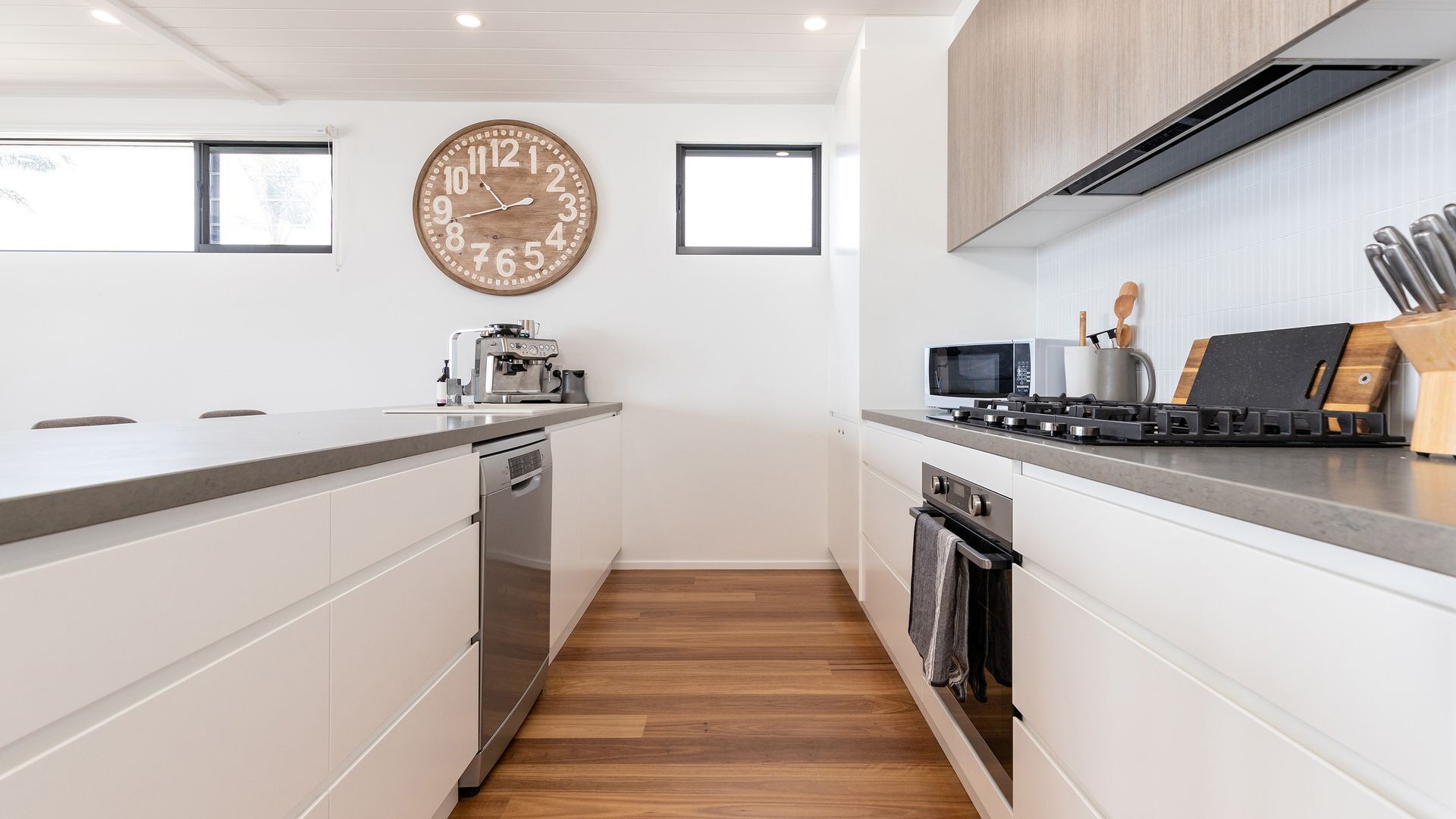 A modern kitchen with white cabinetry, a wooden floor, a large wall clock, and a stove with a microwave above it.
