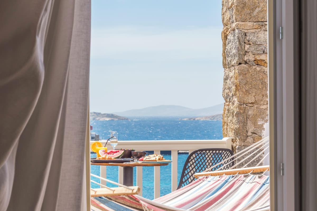 A balcony with a view of the sea, a hammock, and a dining table with food and drinks.