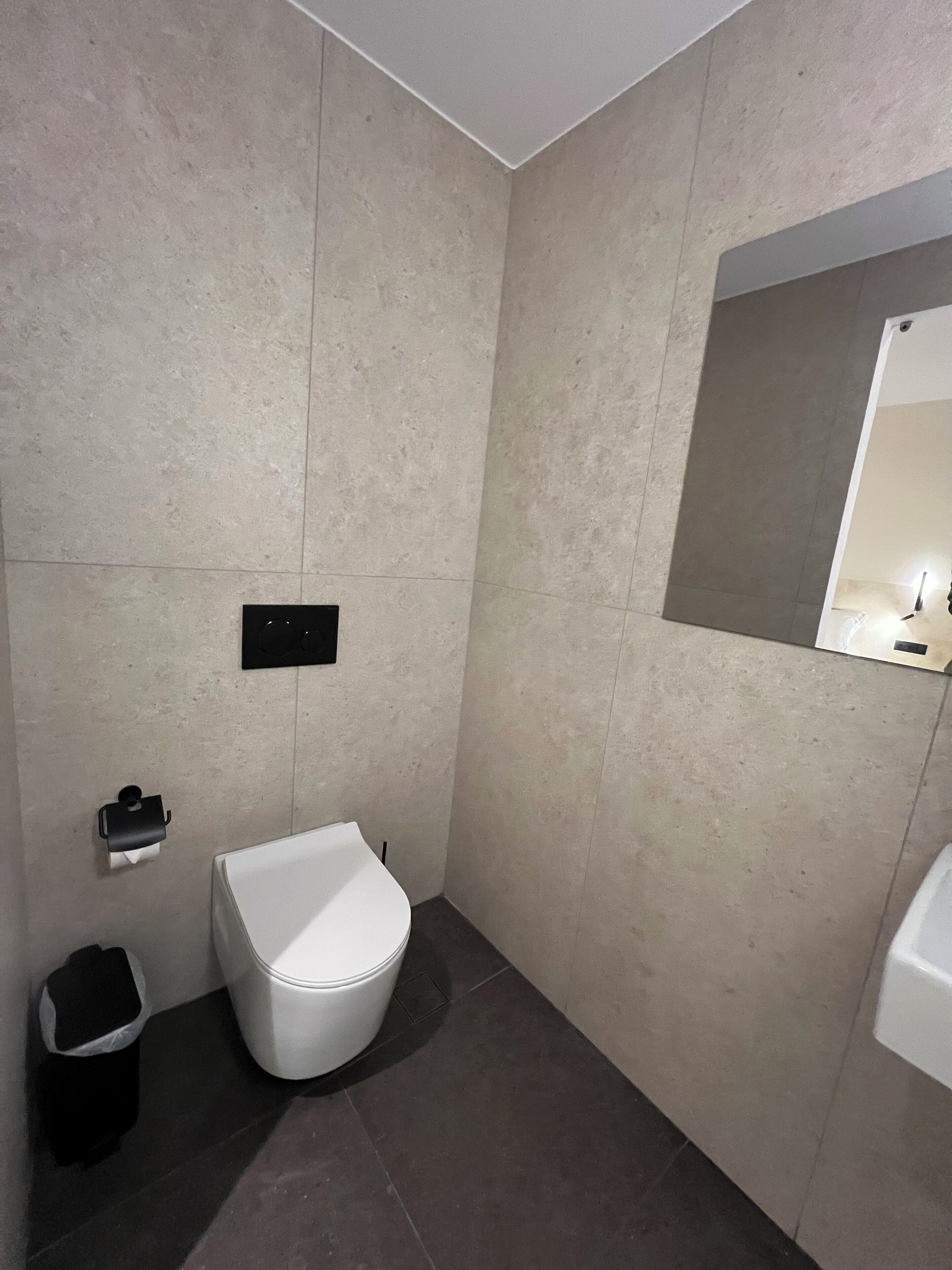 A modern bathroom with a toilet, a trash bin, and a wall-mounted paper towel dispenser.