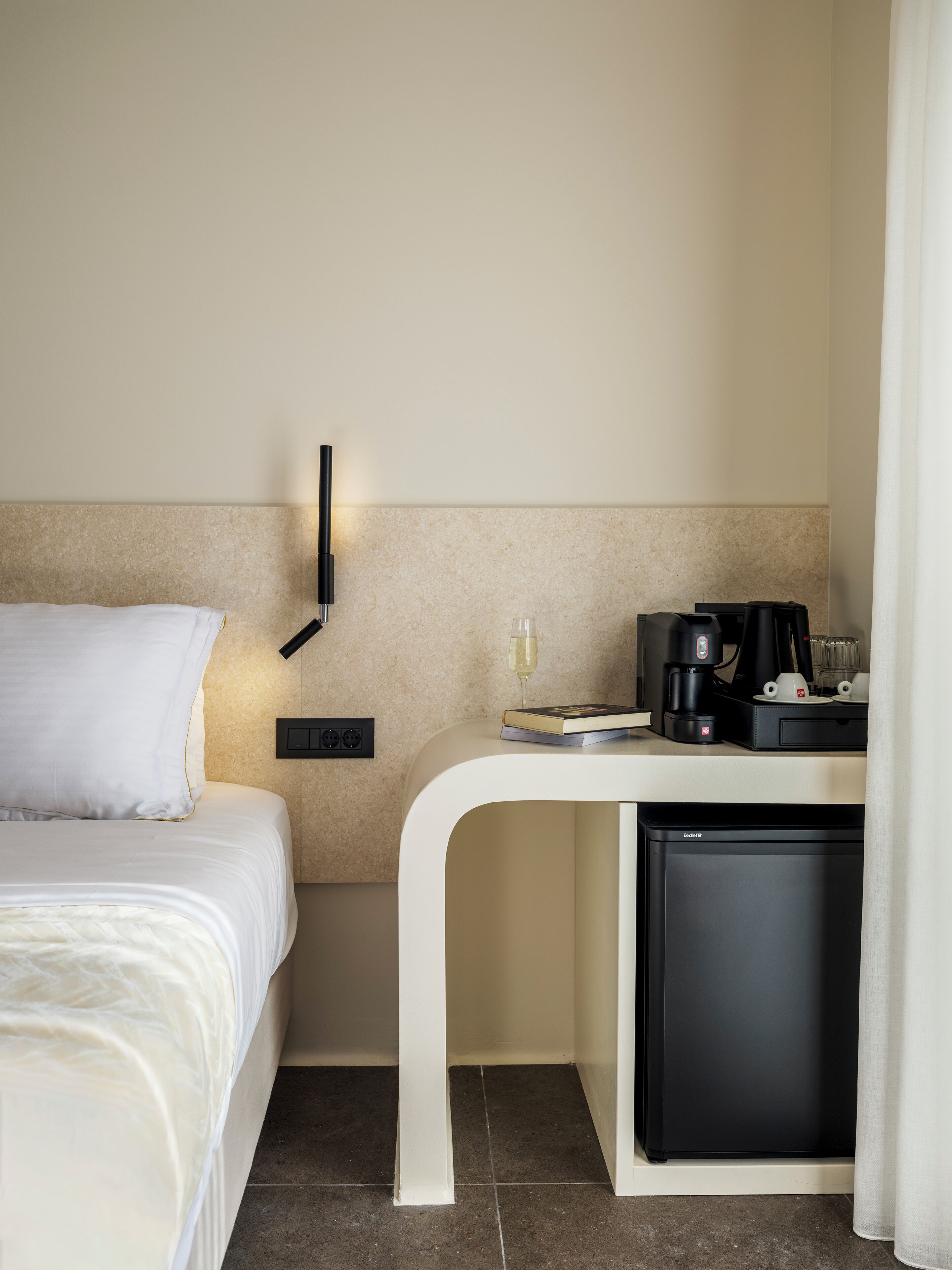 Quarto de hotel com cama, mesa de cabeceira com um livro, máquina de café e um pequeno frigorífico.