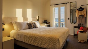 A modern hotel room with a large bed, a desk, a chair, and a view of the city through the window. - Yalos hotel sunset view  (Mykonos)