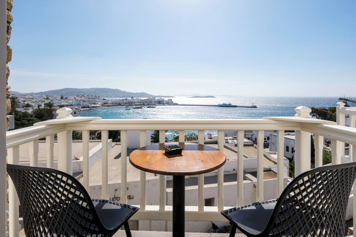 A balcony with two chairs and a small table, overlooking a coastal town with buildings and a body of water.