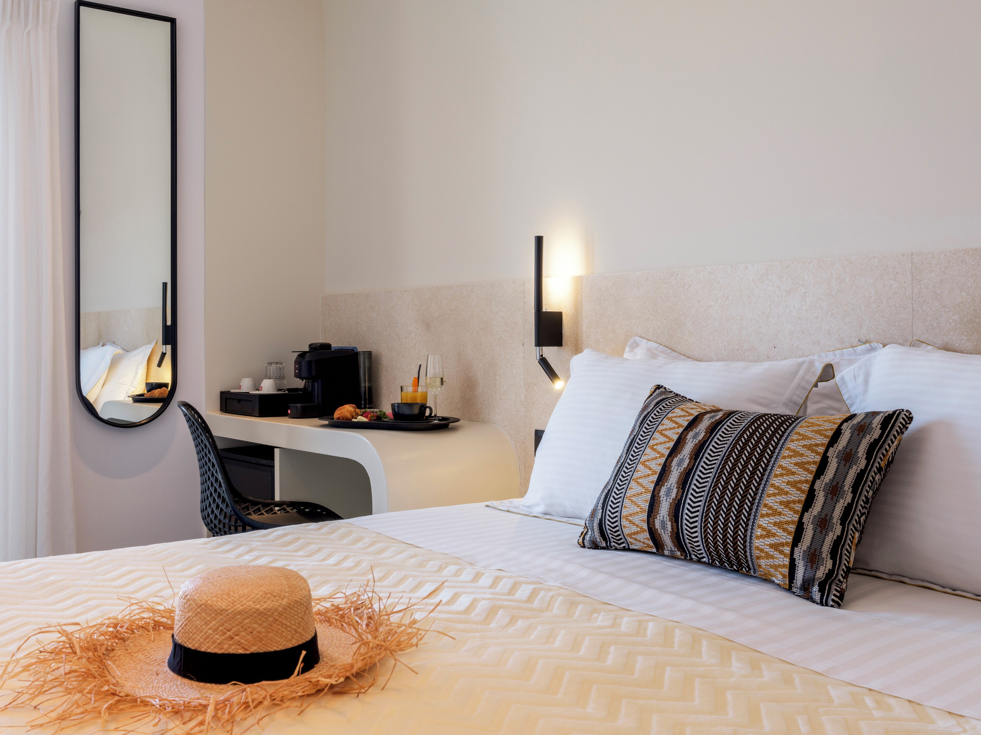 A hotel room with a bed, a desk with a coffee maker, a chair, a mirror, and a wall-mounted light.