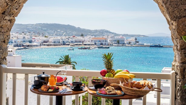 A balcony with a table set for breakfast overlooking a coastal town and the sea. - Yalos hotel sunset view  (Mykonos)