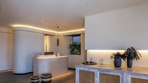 A modern reception area with a curved reception desk, a large mirror, and a minimalist design. There are two woven baskets on the floor and a potted plant on the reception counter. - Yalos hotel sunset view  (Mykonos)