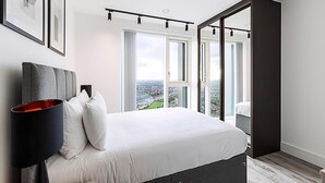 A modern bedroom with a large bed, a black lamp, and a city view through floor-to-ceiling windows.