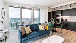 A modern living room with a blue sofa, dining area, and kitchen with dark cabinets and a large window offering a city view.