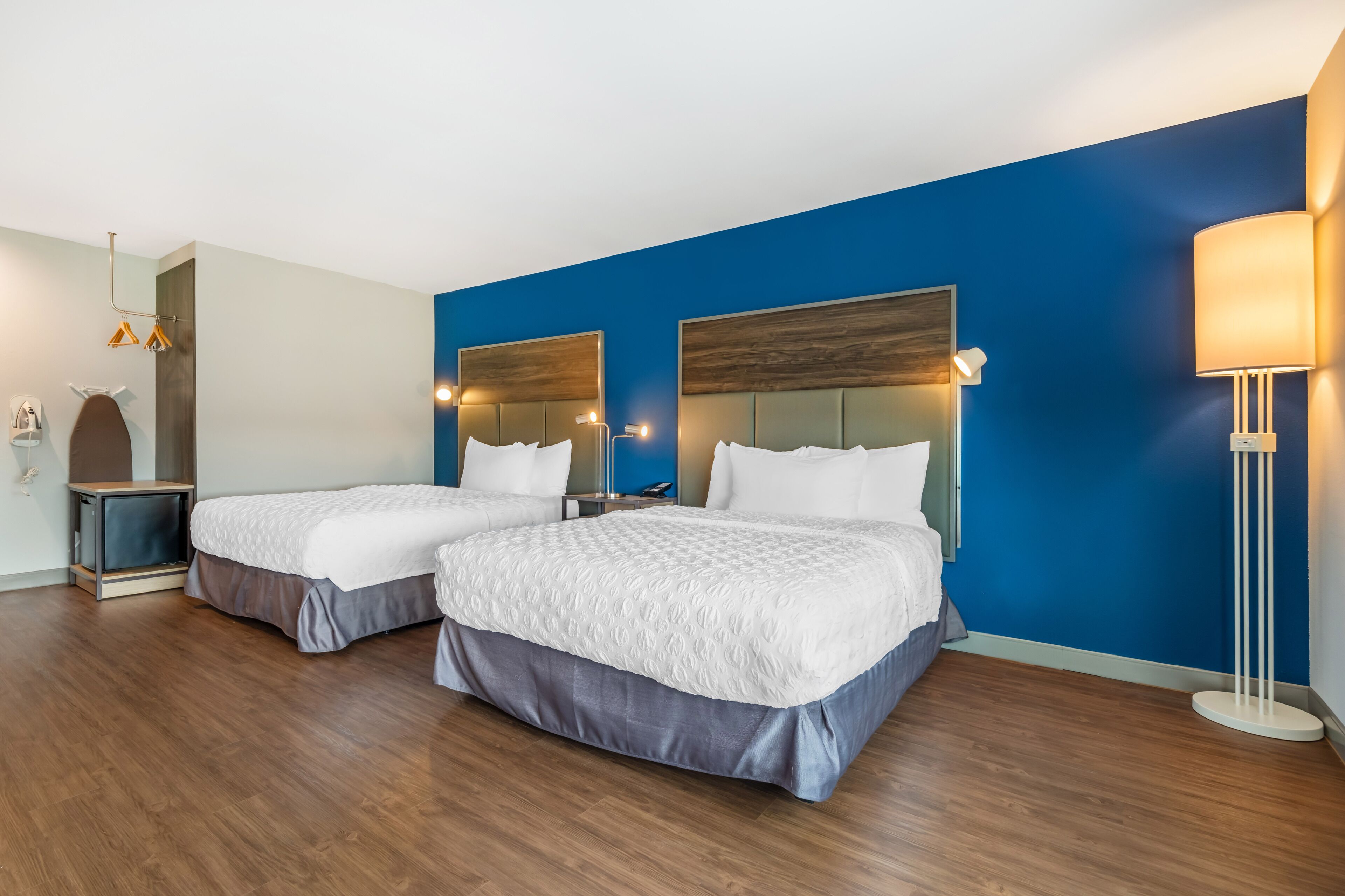 A hotel room with two beds, a blue accent wall, a modern lamp, and a small side table.