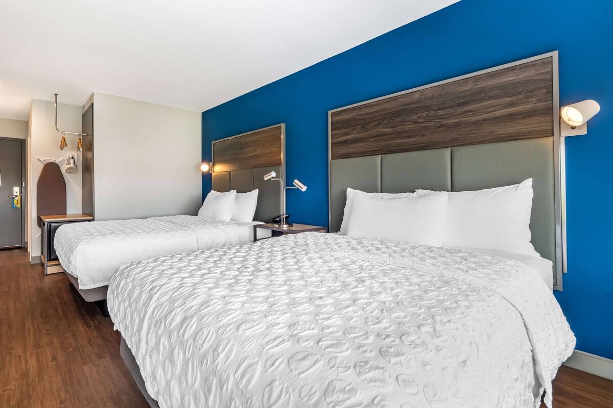 Een hotelkamer met twee bedden, een houten hoofdbord, een blauwe accentmuur en een klein nachtkastje met een lamp.