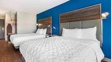 A hotel room with two beds, a wooden headboard, a blue accent wall, and a small bedside table with a lamp.