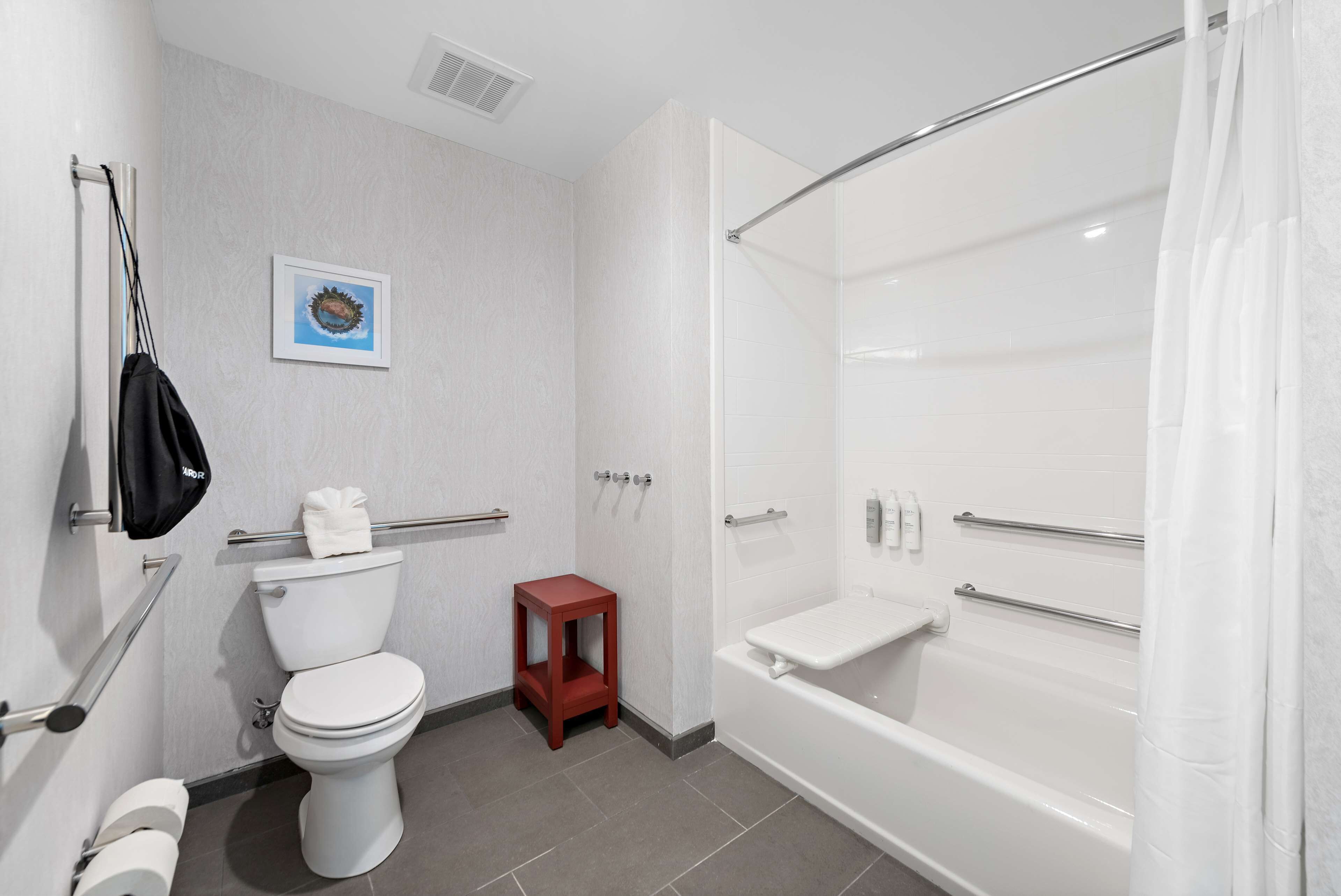 A modern bathroom with a toilet, a small red stool, a bathtub with a shower curtain, and a handrail.