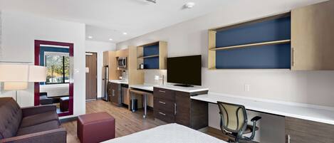Habitación de hotel moderna con cama, escritorio, silla, televisor y estanterías integradas.