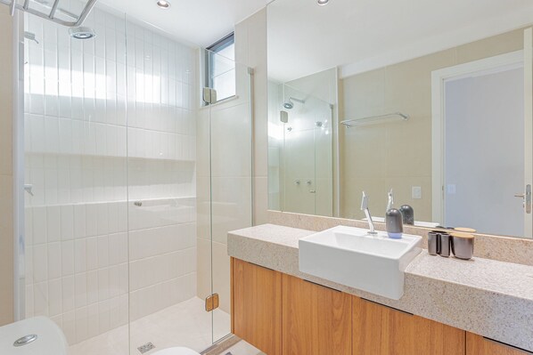 Un baño moderno con ducha envuelta en vidrio, un lavabo blanco y un mueble de tocador de madera.