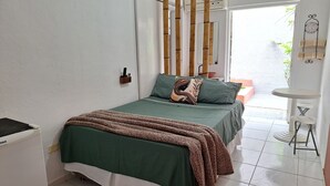 A bedroom with a bed, a bedside table, a small stool, a wall-mounted air conditioner, and a window with a view of greenery outside. - Pousada Oca Poranga (Guarujá)