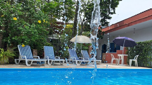 A swimming pool with a water fountain, surrounded by lounge chairs and umbrellas. - Pousada Oca Poranga (Guarujá)