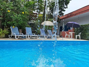 A swimming pool with a water fountain, surrounded by lounge chairs and umbrellas. - Pousada Oca Poranga (Guarujá)