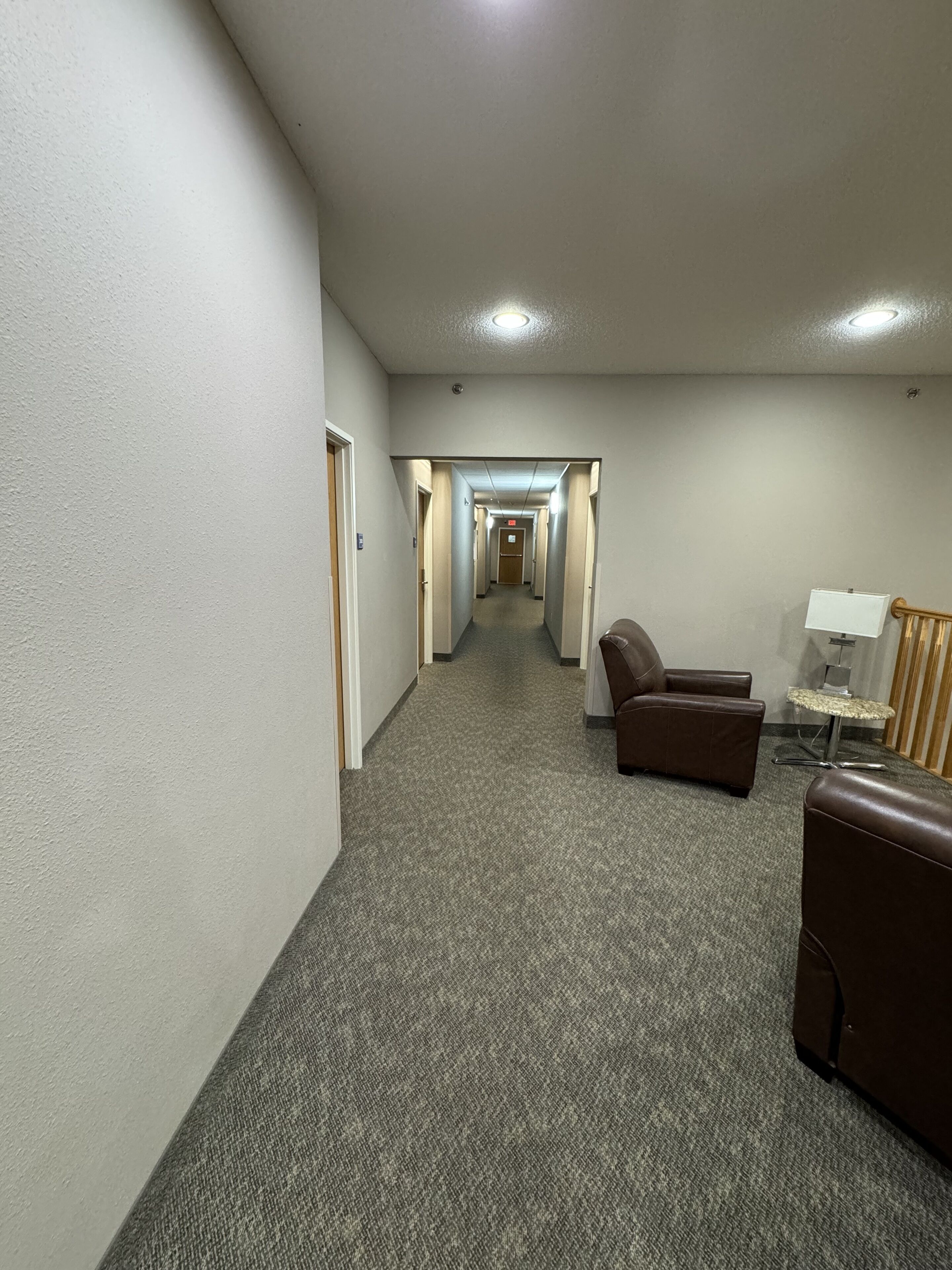 A hotel hallway with carpeted floors, a brown leather sofa, a small table with a lamp, and a cat tree.