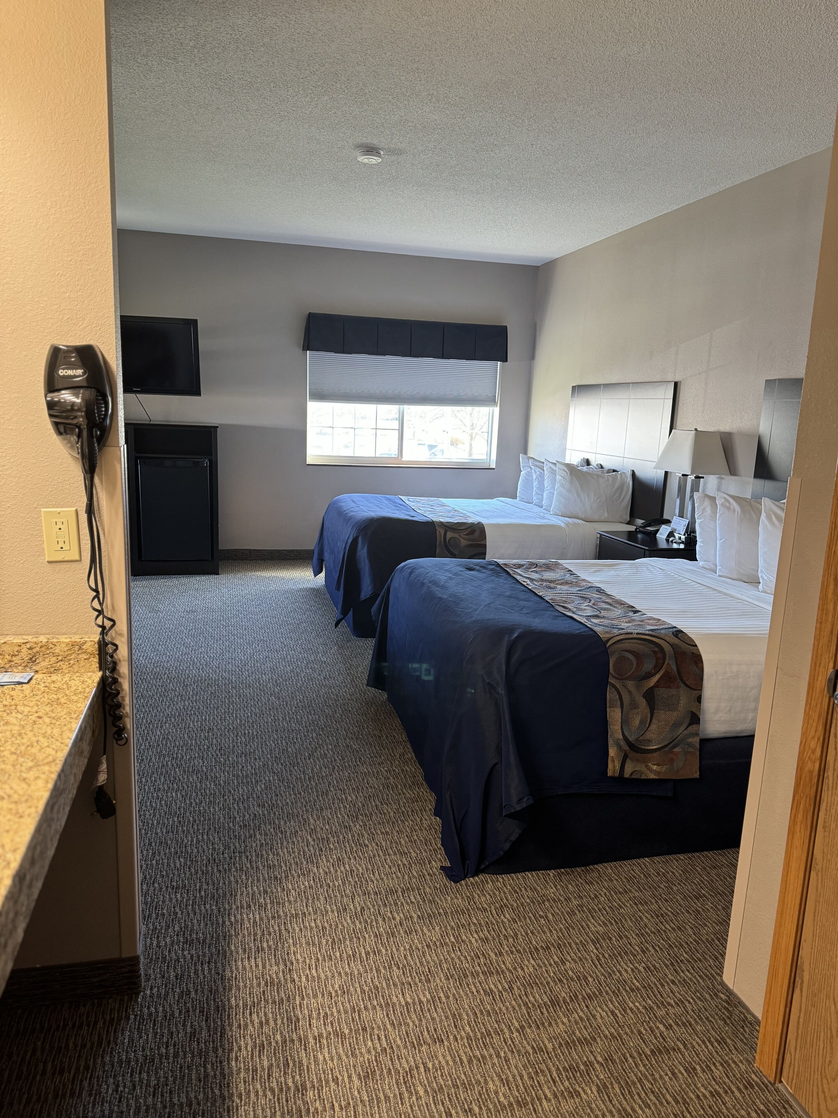 A hotel room with two beds, a desk, a TV, and a window with blinds.