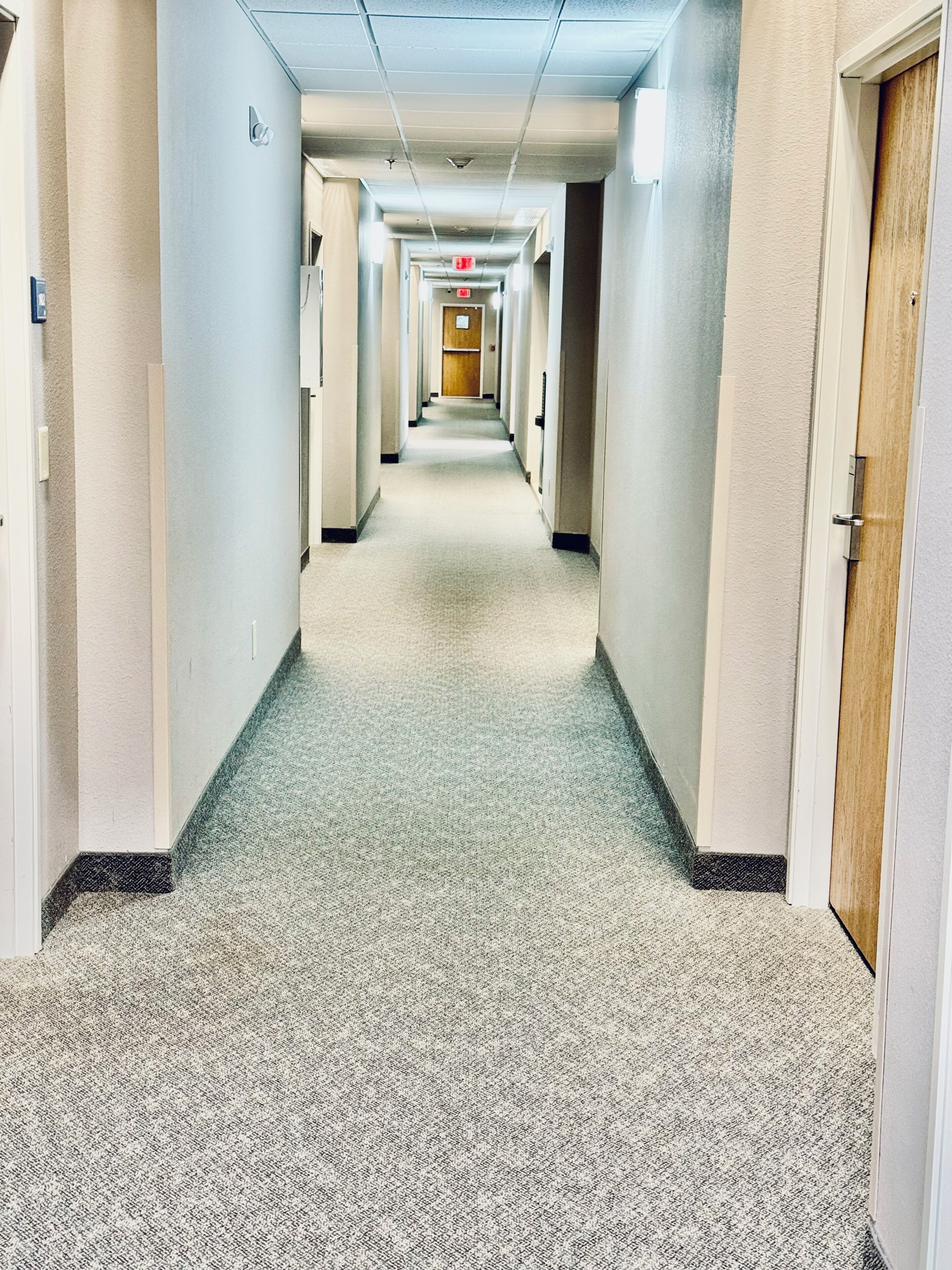 A hotel corridor with carpeted floors, doors on both sides, and a fire exit sign at the end.