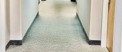A hotel corridor with carpeted floors, doors on both sides, and a fire exit sign at the end.