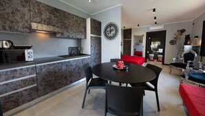 A modern kitchen with a built-in stove, microwave, and kettle. There is a dining area with a round table and black chairs. The room has a red sofa and a decorative wall clock. - One-room Apartment "Suite Love" with View (SCARLINO)
