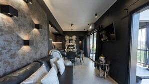 A modern living room with a black leather sofa, a dining area with a round table, and a large window offering a view. - Passion Suite with Garden and Whirlpool (SCARLINO)