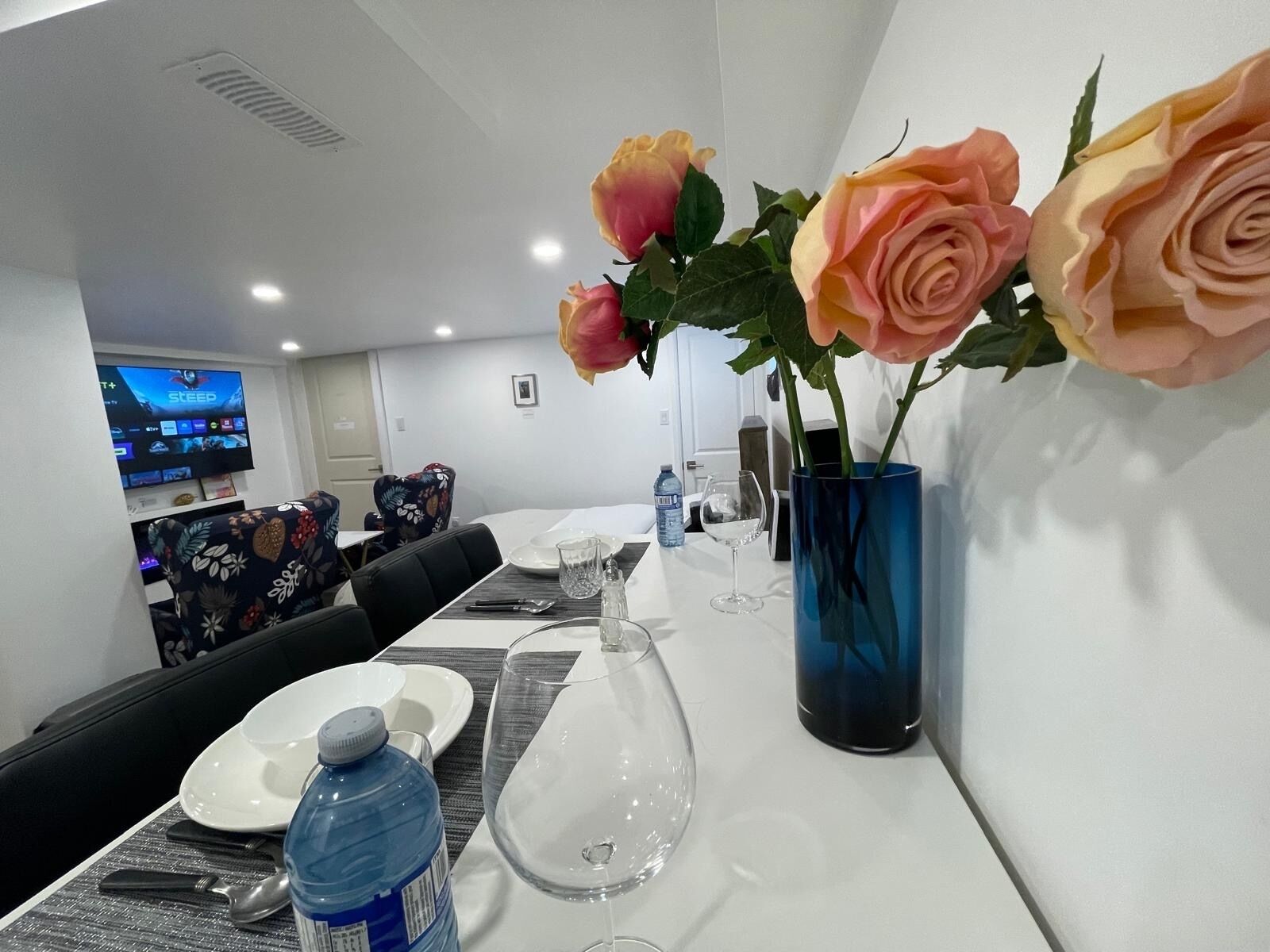 Zona de comedor de hotel con una mesa larga dispuesta para dos, un jarrón azul con rosas y un televisor mostrando un programa de concursos.