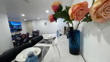 Zona de comedor de hotel con una mesa larga dispuesta para dos, un jarrón azul con rosas y un televisor mostrando un programa de concursos.