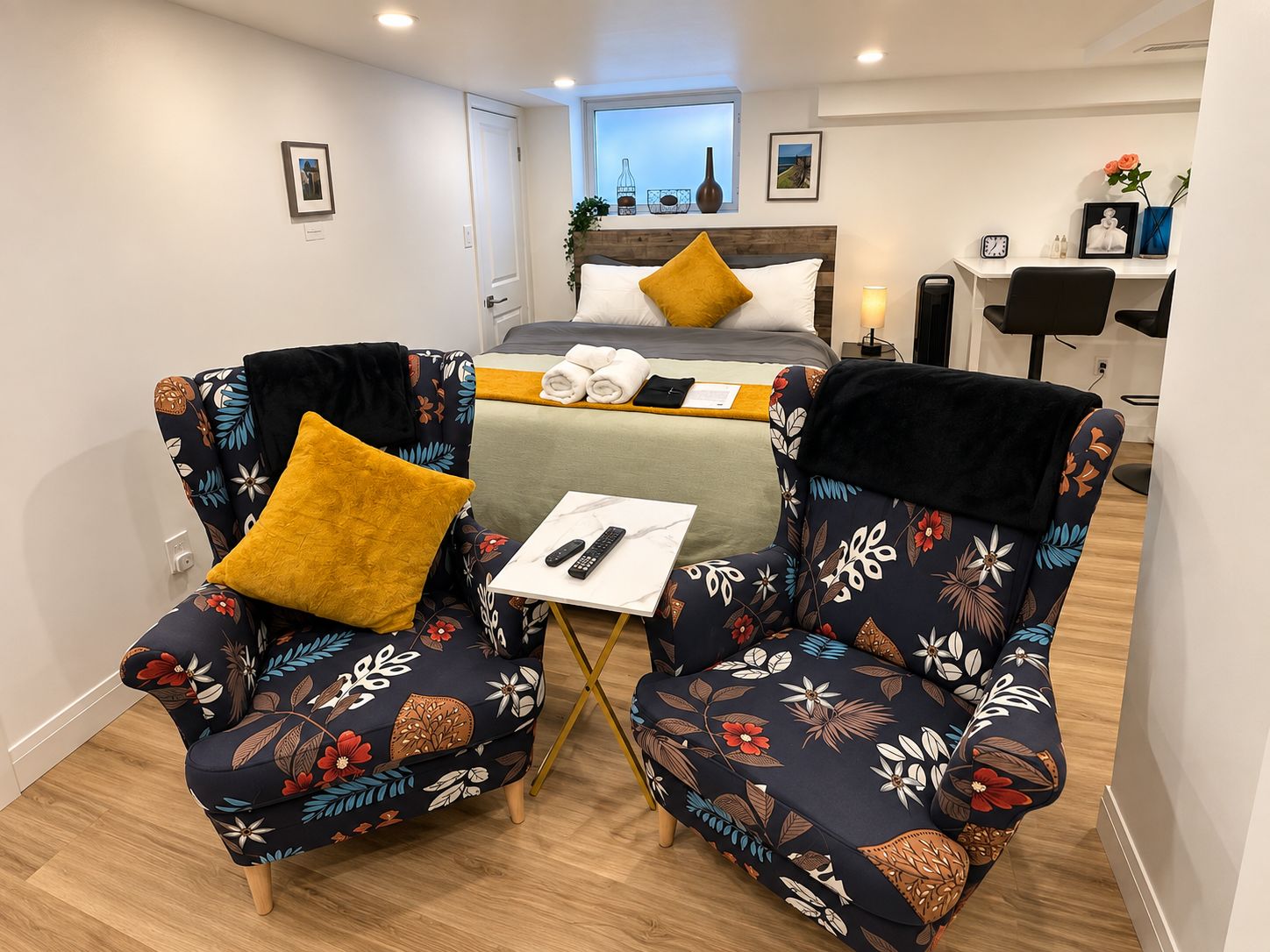 A hotel room with a bed, two armchairs with floral upholstery, a small table with a remote control, and a nightstand with a lamp.