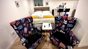 A hotel room with a bed, two floral-patterned armchairs, a small table, and a desk with a chair.