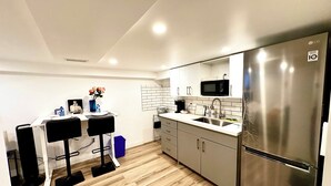 A modern kitchen with a stainless steel refrigerator, a microwave, and a sink. There are two bar stools and a small table with a vase of flowers.