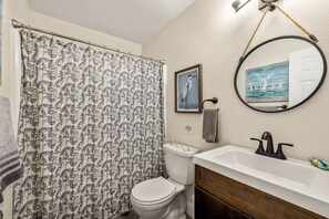 Une salle de bain avec un rideau de douche orné de motifs de feuilles, des toilettes, un lavabo avec un miroir au-dessus et un tableau encadré accroché au mur.