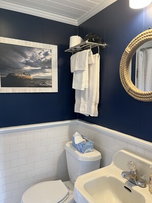 A bathroom with a navy blue wall, a white toilet, a white sink, and a framed picture of a ship on the wall. - Lakeview Cottage, Walk to the beach, parks & downtown. Early booking discount! (Ludington)
