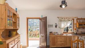 A kitchen with wooden cabinets, a dining table, and a view of a garden through an open door. - Casa Abuela - House in the countryside with pool (Vega de San Mateo)