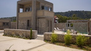 A modern two-story building with a stone facade, a pool area with lounge chairs, and a view of a hilly landscape in the background. - Villa Thoe by Thines Villas (Lefkada)
