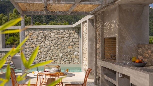 An outdoor patio with a stone wall, a round table with chairs, and a grill area with fruits and glasses. - Villa Thoe by Thines Villas (Lefkada)
