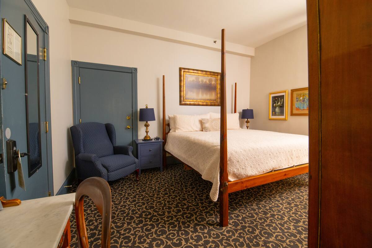 A four-poster bed with a white bedspread, a blue armchair, a nightstand with a lamp, and a patterned carpet.