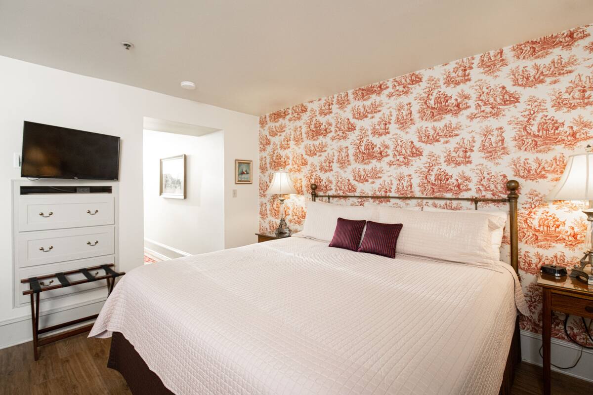 A neatly arranged bedroom with a large bed, a television mounted on the wall, a dresser, and a nightstand with a lamp.