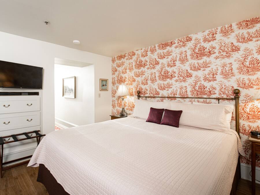 A neatly arranged bedroom with a large bed, a television mounted on the wall, a dresser, and a nightstand with a lamp.