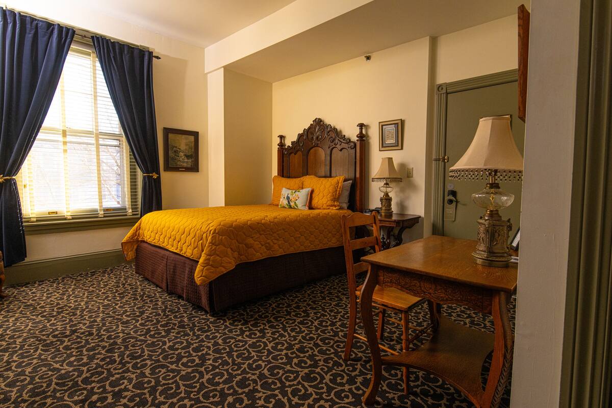 A hotel room with a bed, a desk, a chair, a lamp, and a window with curtains.