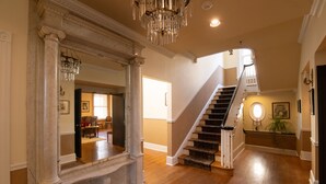 A grand foyer with a marble column, a chandelier, a wooden staircase, and a well-lit living area in the background. - The Lafayette Inn (Easton)