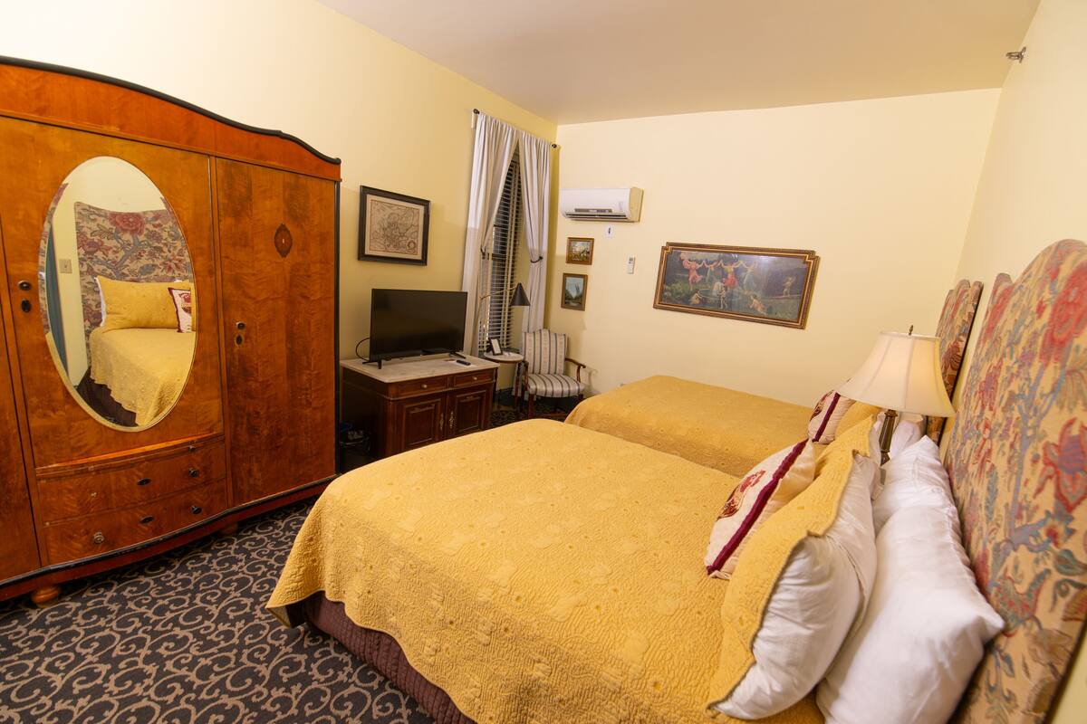 A hotel room with a wooden wardrobe, a television, two beds with yellow bedding, a patterned carpet, and a window with curtains.
