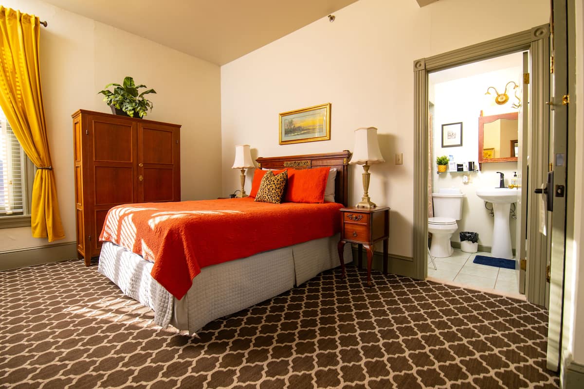 A bedroom with a bed, a nightstand, a dresser, a window with curtains, and a bathroom visible through an open door.