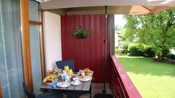 An outdoor dining area with a table set for breakfast, including fruits, bread, and beverages, under a large umbrella.