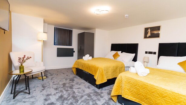 A hotel room with two beds, a sofa, a small table with a vase, and a window with curtains. - The Oasis Apartment (Leeds)